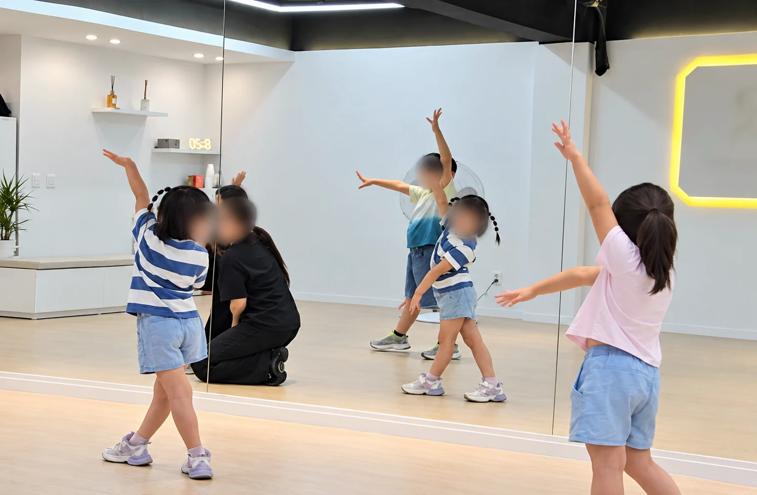 연습실 거울 앞에서 팔을 펼쳐 동작을 익히는 아이들과, 무릎을 꿇고 아이와 눈높이를 맞춘 강사 — 학원 블로그 사진을 AI로 보정, 얼굴은 흐림 처리